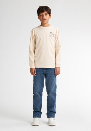 Beige Langarm-T-Shirt mit Brustdruck, kombiniert mit blauen Jeans und weißen Sneakers. Einfaches Design, normale Passform.