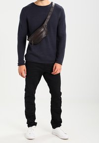 Marinefarbener Strickpullover, schwarze Slim-Fit-Hose und weiße Sneaker. Braune Umhängetasche mit Reißverschluss und verstellbarem Riemen, quer über den Körper getragen.