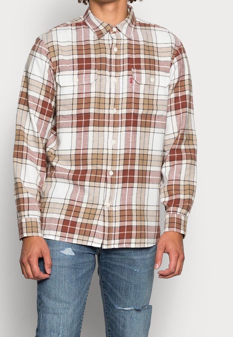 Homme portant une chemise à carreaux marron, beige et blanche avec boutons, deux poches poitrine et un jean bleu délavé.