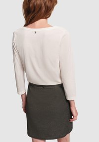 Donna dai capelli rossi che indossa una maglietta bianca a maniche lunghe e una gonna grigia scura, vista di spalle su uno sfondo neutro.