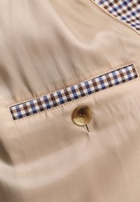 Nærbillede af beige stof med en rektangulær lomme med et ternet mønster i blå, brun og hvid, samt en rund brun knap.