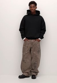 Svart luvtröja, beige mönstrade vida byxor och svarta skor. Luvtröjan har en framficka; byxorna har intrikat sömnad.