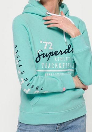 Osoba nosząca turkusową bluzę z kapturem z białymi sznurkami, z napisem „Superdry Athletics Track & Field” oraz jasnoniebieskie dżinsy.