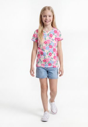 Jente med langt blond hår som smiler, iført en rosa kortermet blomstret skjorte, lyseblå denimshorts og hvite joggesko, går framover.