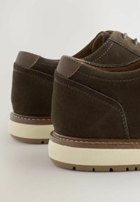 Zapatos de ante marrón con un acento de cuero suave en el talón, que cuentan con una suela de goma beige y detalles de costura en contraste.