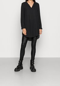 Camisa negra de manga larga con cuello puntiagudo, botones en la parte frontal y dobladillo curvado, combinada con leggings de imitación de cuero negro brillante y botas negras de suela gruesa.