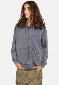 Grå zip-up hættetrøje med snørrebånd, frontlommer og en struktureret overflade. Inkluderer et lille lædermærke med branding på venstre lomme.