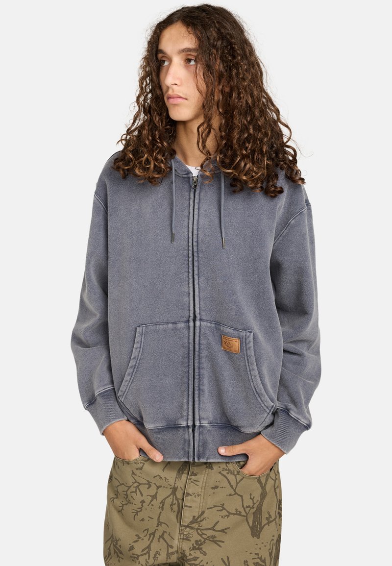Grå zip-up hættetrøje med snørrebånd, frontlommer og en struktureret overflade. Inkluderer et lille lædermærke med branding på venstre lomme.