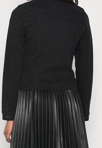 Giacca in denim nera con un design aderente, maniche lunghe e polsini con bottoni, indossata sopra una gonna plissettata in pelle nera.