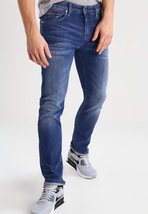 Man draagt vervaagde blauwe spijkerbroek en witte Nike-sneakers, staand tegen een effen lichte achtergrond.
