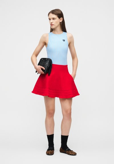 Jeune femme portant un haut sans manches rayé bleu, une jupe rouge, des chaussettes noires, des ballerines imprimé léopard, tenant un sac à franges noir, debout devant un fond blanc.