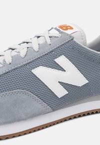 Scarpe sportive grigie con tomaia in mesh, dettagli in suede e logo "N" in pelle bianca. Presentano lacci bianchi e una suola in gomma marrone.