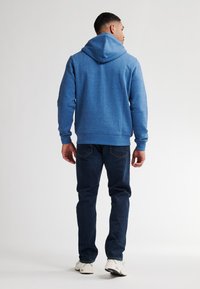 Felpa blu con cappuccio e finitura testurizzata, abbinata a jeans scuri e sneakers bianche. Focus sulla vista posteriore e sui dettagli del cappuccio.