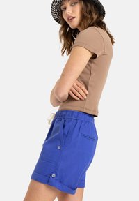 Ljusbrun åtsittande t-shirt tillsammans med blå shorts med dragsko i midjan, uppvikta ärmslut och knappdetaljer.