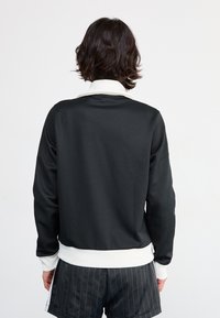 Svart sweatshirt med vit krage och manschetter, slät textur, avslappnad passform, bärs över randiga shorts, visar bakvyn.
