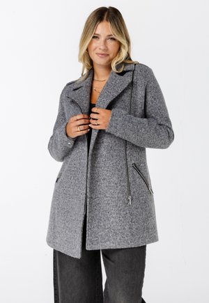 Vrouw die haar grijze wollen jas met zwarte ritsdetails aanpast, gekleed in donkere broeken, gouden ringen en gelaagde colliers, lachend tegen een witte achtergrond.