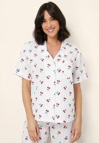 Korte mouwen pyjamashirt van lichtgewicht stof, met een witte basis en een kersenprint in rood en groen. Het heeft een borstzakje en knopen.