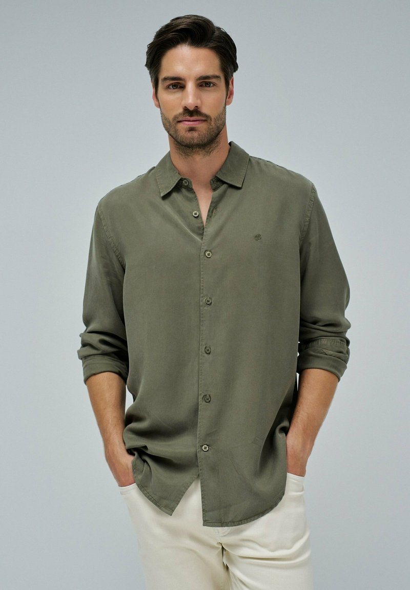 Olivno zelena, srajca z dolgimi rokavi in gumbi iz lahkotnega materiala s priložnostnim ovratnikom in zvitimi rokavi, v kombinaciji s svetlimi hlačami.