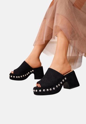 Sabots en suède noir à bout ouvert avec un épais talon bloc, ornés de clous en argent le long de la semelle. Texture douce et silhouette moderne.