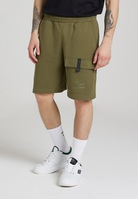 Shorts cargo de culoare verde olive, cu talie elastică, buzunar lateral și logo embosat. Purtați cu pantofi sport albi și șosete albe cu model.