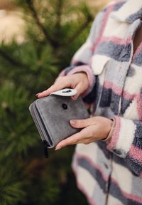 Gray wallet made of textured material with a zipper closure. It has a black button and a white attachment feature. Presented in a hand against a green background.
