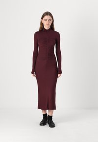 Burgunderfarget tettsittende kjole med lange ermer, høy hals og rynkedetaljer nedover fronten. Stoffet er glatt med en lett glans.
