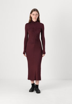Burgunderfarget tettsittende kjole med lange ermer, høy hals og rynkedetaljer nedover fronten. Stoffet er glatt med en lett glans.