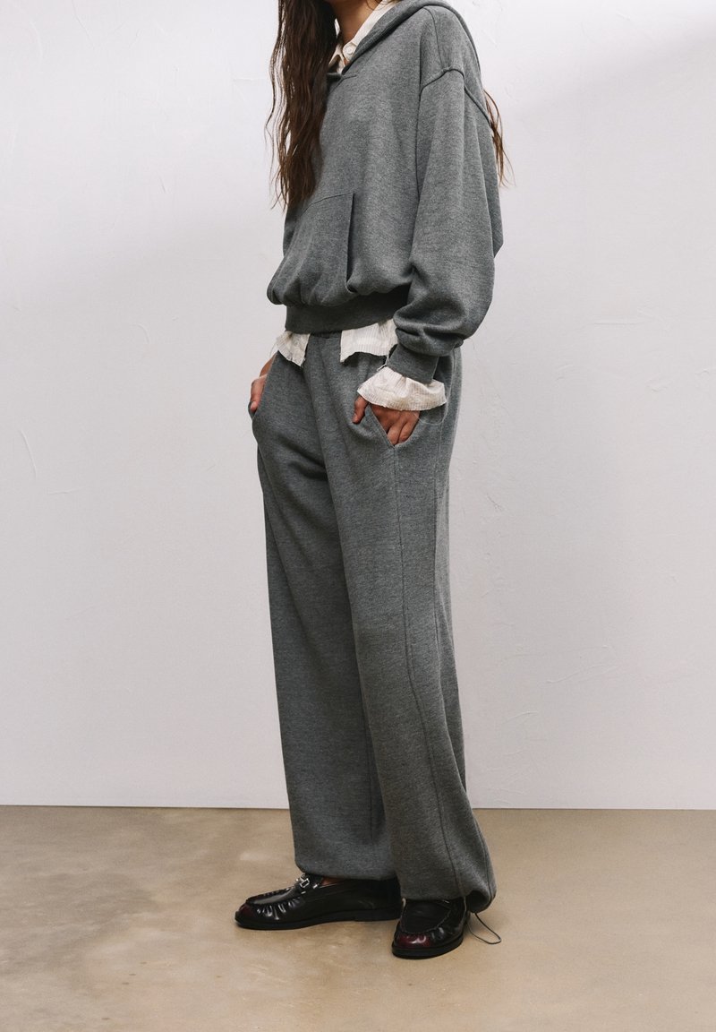 Personne debout portant un sweat à capuche gris oversize avec un pantalon large assorti et des mocassins noirs sur un sol beige contre un mur uni.