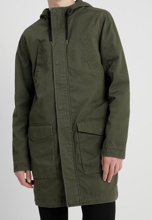 Personne portant une longue veste à capuche vert olive avec deux grandes poches à rabat à l'avant et deux poches poitrine, debout devant un fond uni.