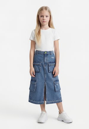 Młoda dziewczyna z długimi blond włosami, ubrana w białą koszulkę z krótkim rękawem, niebieską jeansową spódnicę cargo i białe trampki, stojąca na jednolitym tle.