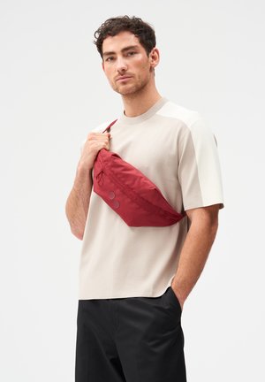 Mann mit kurzen lockigen Haaren, trägt beige Kurzarmoberteil und schwarze Hose, hält eine rote Umhängetasche quer über der Brust, steht vor einfarbigem Hintergrund.