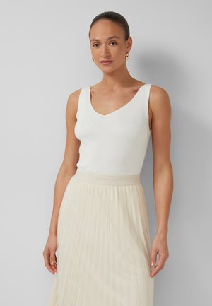 Kobieta w białym, bezrękawnym topie z dekoltem w serek i kremowej plisowanej spódnicy, stojąca na tle jednolitego szarego tła.