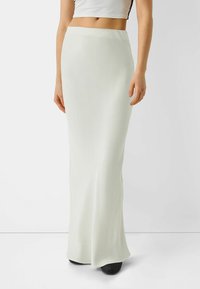 Žena oblečená v krémové maxi sukni s vysokým pasem, kterou doplňuje o černé strukturované boty, na pozadí čistě bílé.