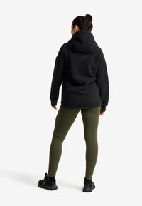 Svart fleecetröja med stor huva, ribbade ärmmuddar och en diskret logga på ryggen. Matchas med åtsittande olivgröna leggings och svarta sneakers.