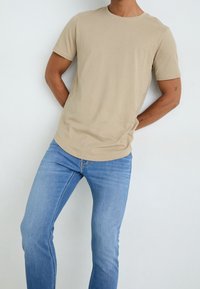 Béžové bavlněné tričko s kulatým výstřihem, v kombinaci s světle modrými džínovými kalhotami. Tričko má krátké rukávy a volnější střih.