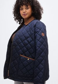 Μπουφάν με επένδυση σε μπλε ναυτικό χρώμα, με ταμπά γιακά, χρυσά μεταλλικά στοιχεία και πλαϊνή τσέπη με καφέ περίγραμμα και έμβλημα, σχεδιασμένο για άνετη εφαρμογή.