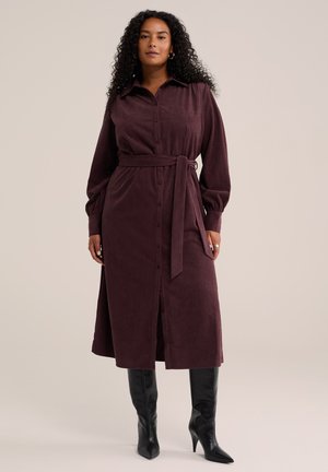 Femme portant une robe en velours côtelé violet foncé à manches longues, boutonnée, avec une ceinture à la taille et des bottes noires à talons montant jusqu'aux genoux.