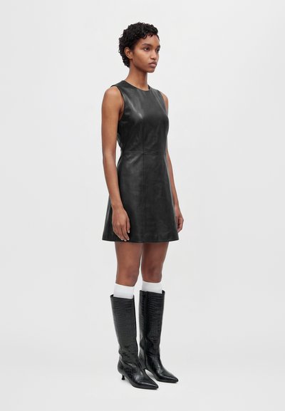 Un mannequin se tient devant un fond gris clair, portant une robe noire sans manches, des chaussettes blanches et des bottes noires montant jusqu’aux genoux.