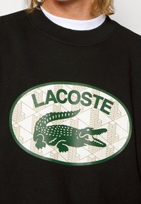 Sudadera negra con un gran logo ovalado que presenta un cocodrilo verde y el texto "LACOSTE", sobre un fondo texturizado color crema.