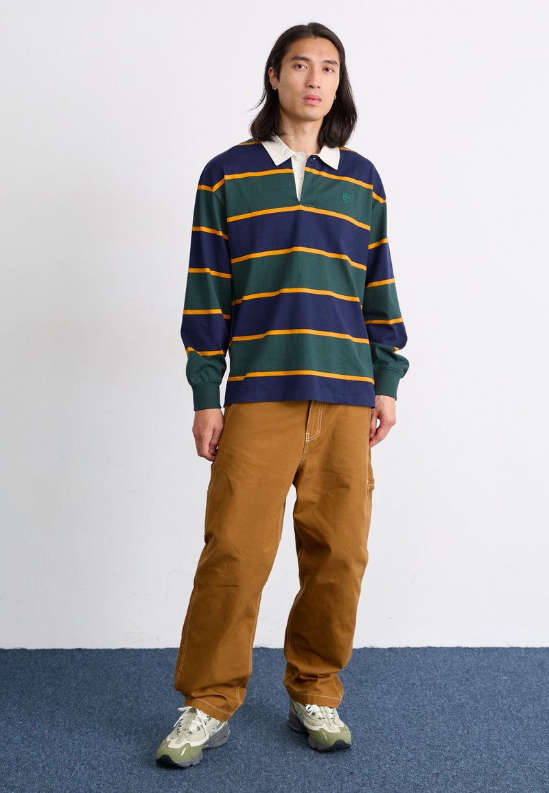 Polo de manga larga a rayas en navy, verde y naranja; cuenta con un cuello blanco en contraste y un logo. Combinado con pantalones sueltos de color beige.
