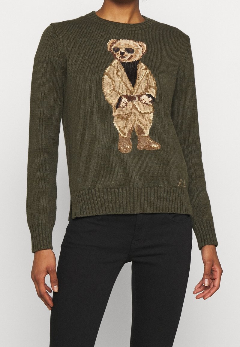 Pull en laine vert olive avec un ours brodé au centre portant des lunettes de soleil, une légère veste et un haut sombre. Bord-côte aux poignets et à l'ourlet.