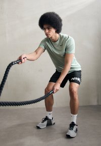 Uomo atletico con una maglia Nike verde chiaro e pantaloni corti neri che utilizza una corda da battaglia nera. Indossa scarpe Nike grigie e nere con calzini bianchi.
