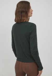 Tamnozeleni pleteni džemper s dugim rukavima, okruglim izrezom i nabranim manžetama. Tkanina izgleda glatko i lagano, s krojem koji ističe figuru.