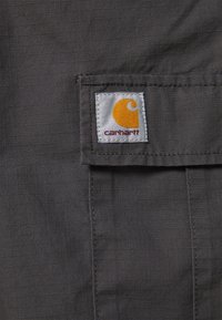 Grauer Stoff mit Karomuster, versehen mit einem angenähten Carhartt-Logopatch in Orange und Weiß, der an der Kante einer Tasche befestigt ist.