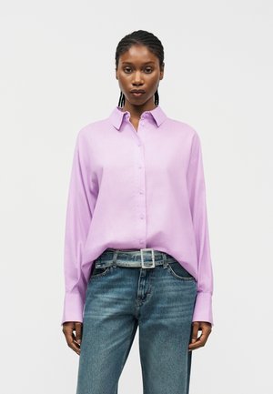 Γυναίκα με πλεξούδες, φορώντας ένα μωβ πουκάμισο με κουμπιά και μακριά μανίκια, το οποίο είναι μπα tucked σε ψηλόμεσα μπλε τζιν με γκρι ζώνη.