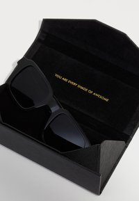 Occhiali da sole neri con forma rettangolare, finitura opaca, contenuti in un astuccio di tessuto nero. All'interno il testo recita "SEI OGNI SFUMATURA DI FANTASTICO."