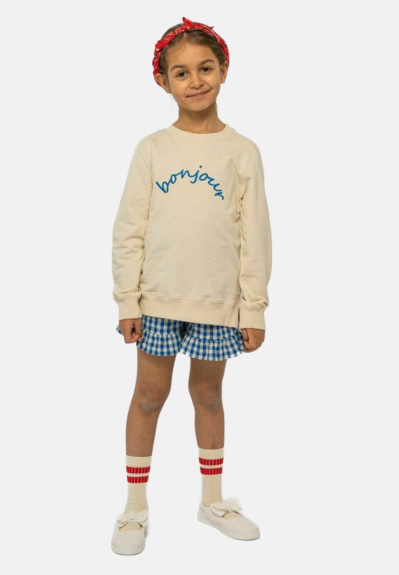 Krämfärgad sweatshirt med "bonjour" i blått, rutiga blå shorts, vita skor med rosetter och rödrandiga beige strumpor. Röd bandana i håret.