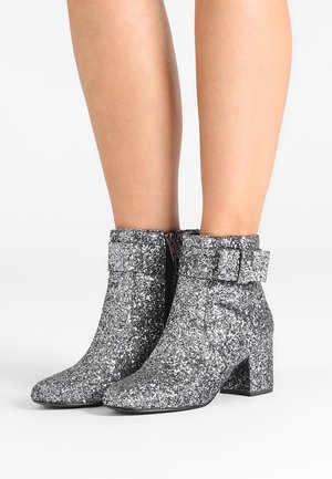 Enkellange zilveren glitterlaarzen met blokhakken en zijgespen, gedragen op blote benen tegen een witte achtergrond.