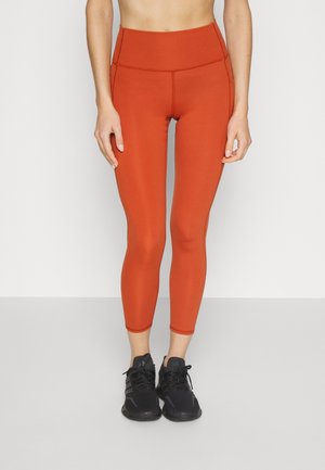 Leggings taille haute et ajustées en orange rouille, fabriquées en tissu extensible avec des coutures plates. Assortis avec des chaussures de sport noires.