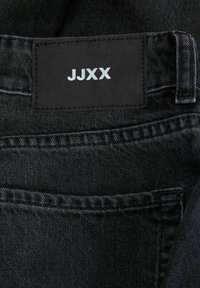 Vaqueros de mezclilla negra con una tela texturizada, un bolsillo trasero visible y una etiqueta rectangular negra con el texto en relieve en blanco "JJXX."